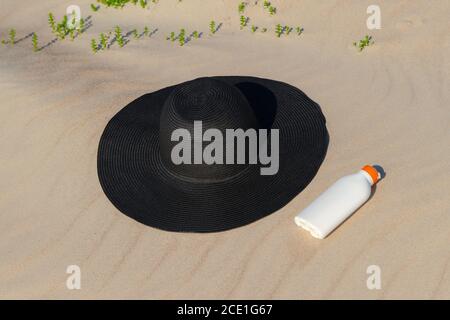 Sonnencreme neben einem schwarzen Hut auf dem Sand an einem sonnigen Tag am Strand. Kin Schutz Kosmetik zum Sonnenbaden. Niemand Stockfoto