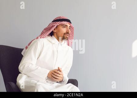 Der seriöse arabische Mann sieht sitzend aus, während er auf einem Sessel über grauem Hintergrund sitzt Stockfoto