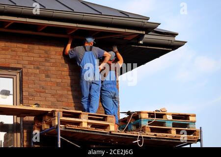 Arbeiter während der Bedachung, zwei Bauarbeiter auf Gerüst stehen in der Nähe der Gebäudewand. Bau- und Reparaturarbeiten eines Hauses Stockfoto