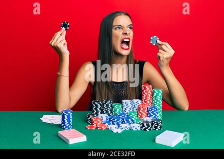 Junge hispanische Frau sitzt auf dem Tisch hält Casino-Chips wütend und verrückt schreien frustriert und wütend, schreien vor Wut. Wut und Aggressiv Stockfoto