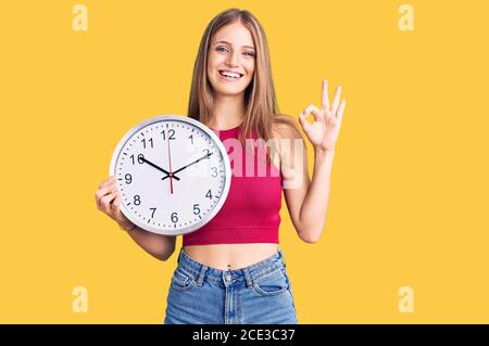 Junge schöne blonde Frau hält große Uhr tun ok Zeichen mit Fingern, lächelnd freundlich gestikulieren ausgezeichnete Symbol Stockfoto