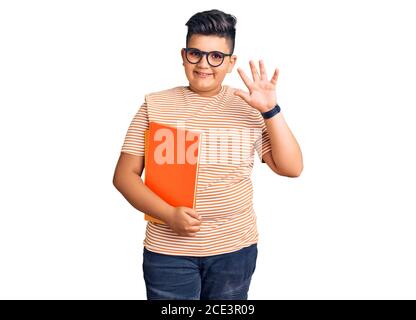 Kleiner Junge Kind hält Buch trägt Brille waiving sagen Hallo glücklich und lächelnd, freundliche Willkommensgeste Stockfoto