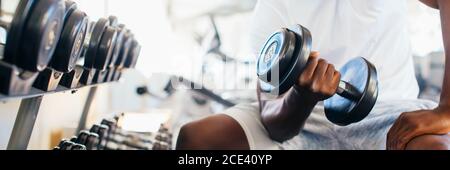 Junge afrikanische amerikanische Mann sitzt und heben eine Hantel in der Nähe des Racks im Fitnessstudio. Männliche Person, ein Krafttraining Bizeps in Fitnesscenter curl Stockfoto