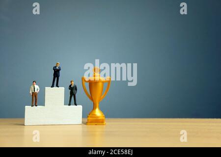 Miniatur-Geschäftskonzept - Geschäftsmann vor Weiß stehen Siegerpodium mit goldener Trophäe Stockfoto