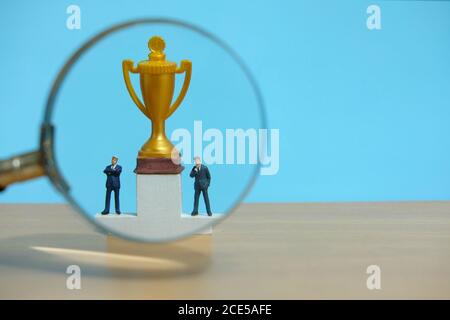 Miniatur-Geschäftskonzept - Geschäftsmann vor Weiß stehen Podium mit goldener Trophäe und Lupenglas Stockfoto