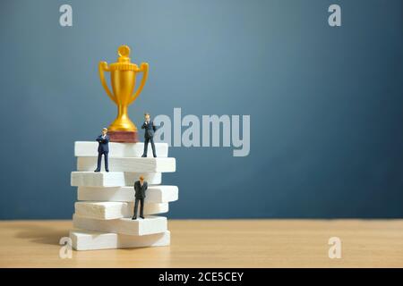 Miniatur Business-Konzept - Geschäftsmann auf weißen Treppe Leiter stehen Mit goldener Trophäe darüber Stockfoto