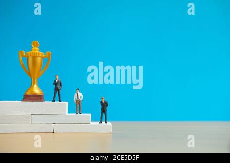 Miniatur-Geschäftskonzept - Geschäftsmann auf weißem Siegerpodest stehen Mit goldener und silberner Trophäe Stockfoto