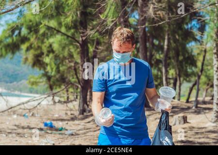 Volunteer Mann in Gesichtsmaske abholen Müll verschmutzen Strand in der Nähe von Meer, halten Blick auf weggeworfene Einweg-Plastikbecher. Problem mit verschüttetem Müll Stockfoto