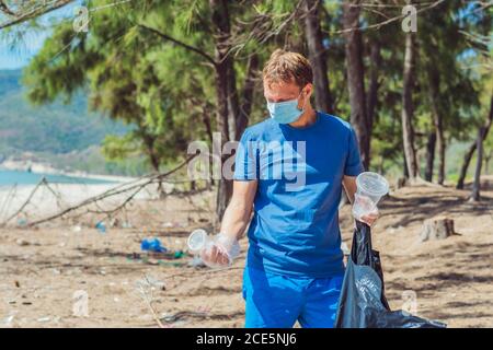 Volunteer Mann in Gesichtsmaske abholen Müll verschmutzen Strand in der Nähe von Meer, halten Blick auf weggeworfene Einweg-Plastikbecher. Problem mit verschüttetem Müll Stockfoto