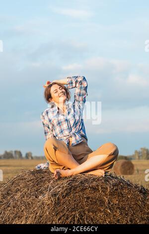 Romantische Brünette Frau in einem karierten Hemd und Hose sitzt auf einem runden Heuhaufen in einem erstaunlichen Feld, im Hintergrund Wald. Warme Farben der Sonnen Stockfoto