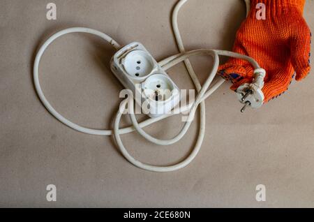 Die Hand trägt einen orangefarbenen Schutzhandschuh und hält nach dem Brand einen elektrischen Stecker. Weiße Steckdosenleiste nach Kurzschluss. Elektrische Sicherheit. Stockfoto