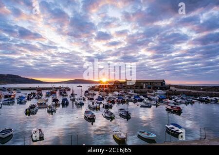 Lyme Regis, Dorset, Großbritannien. August 2020. UK Wetter: Die aufgehende Sonne platzt durch die Wolken bei Sonnenaufgang über dem Cobb bei Lyme Regis an einem schönen, aber kühlen August Bank Holiday Montagmorgen. Die spektakulären Wolken am Himmel spiegeln sich im stillen Wasser des Hafens. Kredit: Celia McMahon/Alamy Live Nachrichten Stockfoto