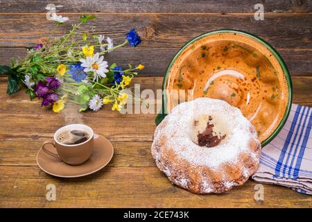 Ein Gugelhupf mit seiner Keramik-Backform und einer Tasse Kaffee und einem Feld Blumen Geburtstagsstrauß auf einem Holztisch mit Holzhintergrund. Stockfoto