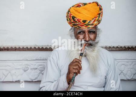 Indien - 30. Oktober 2012: Inhalt älterer indischer Mann in traditionellem Turban sitzt in der Nähe von Steingebäude und raucht Shisha, während er sich entspannt und wegschaut Stockfoto