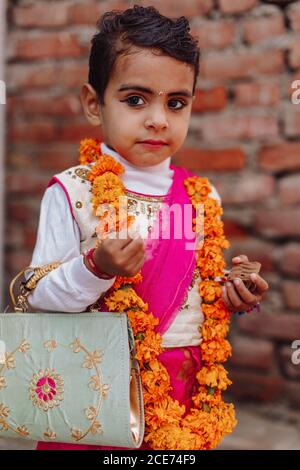 Indien - 9. November 2012: Nettes indisches kleines Mädchen in festlicher stilvoller Kleidung, das auf dem Hintergrund der Ziegelmauer in der Stadt steht und wegschaut Stockfoto