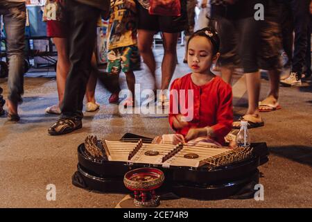 Thailand - 14. August 2010: Ruhiges thailändisches Mädchen, das auf dem Bürgersteig in einer überfüllten Straße sitzt und Melodie auf dem traditionellen khim spielt Stockfoto