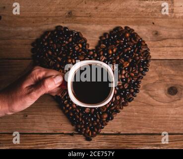 Von oben der anonymen Person mit dem Becher des heißen Getränks Platziert auf Kaffeebohnen in Form von Herz Stockfoto