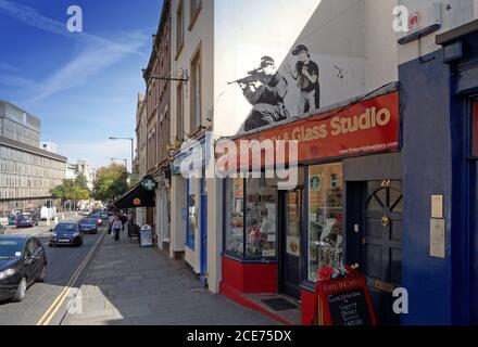 Banksy-Kunstwerk in der Upper Maudlin Street in Bristol eines hockigen bewaffneten Polizisten, mit einem Kind, das kurz vor dem Platzen einer Papiertüte hinter ihm (jetzt weg) Stockfoto