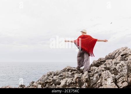 Lange Rückansicht der Frau in stilvoller Kleidung, die am Rand der felsigen Klippe gegen den bewölkten Himmel steht und die Küste bewundert, während Sie Ihren Urlaub an der asturischen Küste Spaniens genießen Stockfoto