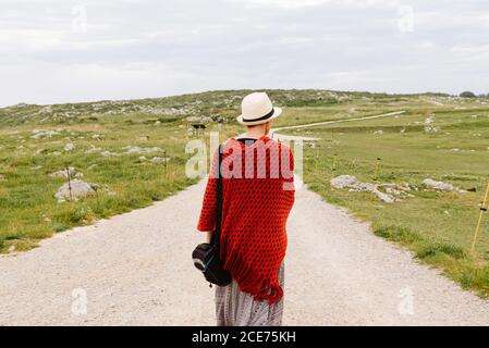 Rückansicht einer nicht erkennbaren weiblichen Reisenden in rot gestricktem Poncho Und Hut zu Fuß auf unbefestigten Weg durch grasbewachsenen Hügeln Genießen Sie Ihren Urlaub in Asturien in Spanien Stockfoto