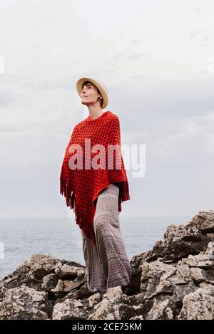 Von unten voller Länge der Frau in stilvoller Kleidung stehen auf dem Rand der felsigen Klippen gegen bewölkten Himmel und bewundern Seeslandschaft während des Urlaubs an der asturischen Küste von Spanien Stockfoto