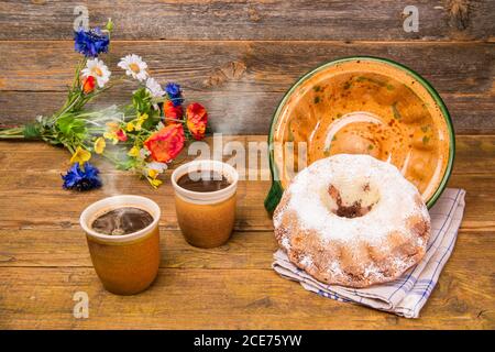 Ein Gugelhupf mit seiner Keramik-Backform und zwei Tassen Kaffee und einem Feld Blumen Geburtstagsstrauß auf einem Holztisch mit Holzhintergrund. Stockfoto