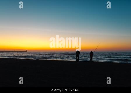 Silhouetten von anonymen Freunden fangen Fische mit Angelrute während Stehen am Ufer des Meeres unter malerischen bunten Himmel bei Sonnenuntergang Stockfoto