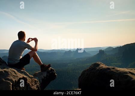 Tourist in grau T-Shirt macht Fotos mit Smartphone auf dem Gipfel des Felsens. Verträumte hügelige Landschaft unten, orange rosa nebliger Sonnenaufgang i Stockfoto