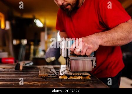 Konzentrierter männlicher Tischler, der an der Werkbank in der Werkstatt und beim Polieren steht Holzbrett mit Bandschleifmaschine Stockfoto