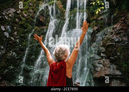 Rückansicht der nicht erkennbaren weiblichen Touristen bewundern majestätische Landschaft Wasserfall und genießen Sie die Freiheit mit erhobenen Armen während der Sommerferien Stockfoto