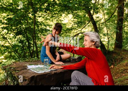 Seitenansicht der jungen und älteren weiblichen Reisenden, die auf dem Platz sitzen Stein im Wald und diskutieren Ort während der Orientierung auf der Karte Und Kompass Stockfoto