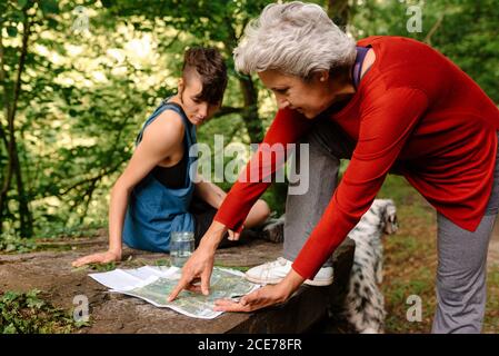 Seitenansicht der jungen und älteren weiblichen Reisenden, die auf dem Platz sitzen Stein im Wald und diskutieren Ort während der Orientierung auf der Karte Und Kompass Stockfoto