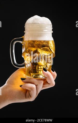 Crop weiblich mit Glas in Form von Schädel gefüllt mit Frisches Bier mit Schaum auf schwarzem Hintergrund im Studio Stockfoto