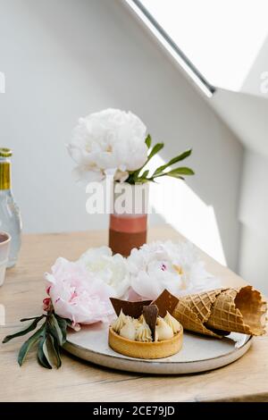 Hoher Winkel von schmackhaften Karamell Tart garniert mit weißen und Milchschokolade und auf Tablett mit Waffelzapfen und Pfingstrosen Stockfoto