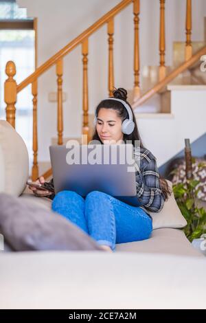 Junge Studentin in Freizeitkleidung mit Kopfhörern beim Video ansehen Unterricht auf dem Laptop, während Sie auf dem Sofa in einer modernen Wohnung sitzen Stockfoto