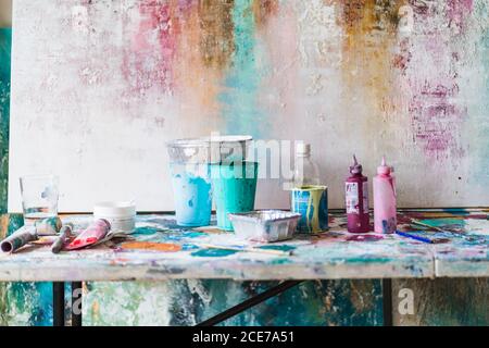 Plastikflaschen mit bunten Farben und leere Behälter in der Nähe Künstler Werkzeuge auf schmutzigem Tisch in der Nähe mehrfarbiger Wand mit unebener Oberfläche Im Kunststudio Stockfoto