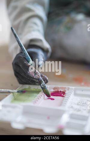 Crop anonyme weibliche Illustratorin in schmutzigen Gewand und Handschuhe sitzen Auf dem Boden mit Pinsel vor der Palette mit Gouache Farben in der Werkstatt Stockfoto