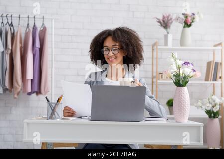 afroamerikanische Frau prüft Dokumente, trinkt Kaffee, sitzt am Tisch Stockfoto