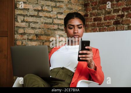 Fokussierte, elegante afroamerikanische Frau, die Nachrichten auf dem Mobiltelefon abcheckt, während sie mit einem Laptop im Sessel gegen eine Backsteinwand in einer Loft-Wohnung sitzt Stockfoto