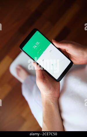 Von oben Ernte anonyme Weibchen in Pyjama stehend auf hölzern Boden in der Nähe der intelligenten digitalen Waage und mit BMI-Steuerung Anwendung auf Handy mit Touchscreen Stockfoto