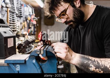 Seitenansicht des konzentrierten männlichen Mechanikers mit Tattoos mit Schraubstock Und die Reparatur der roten Rückleuchte des Motorrads während der Arbeit in Werkstatt Stockfoto