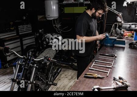 Seitenansicht des konzentrierten männlichen Mechanikers mit Tattoos mit Schraubstock Und die Reparatur der roten Rückleuchte des Motorrads während der Arbeit in Werkstatt Stockfoto