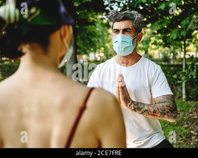 Tätowierten Mann mittleren Alters in Schutzmaske, der mit namastiger Geste vor einer nicht erkennbaren Frau in der Göttin Asana steht, während er gemeinsam im Sommerpark Yoga praktiziert Stockfoto