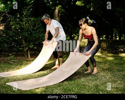 Vollkörper von erwachsenen bärtigen männlichen und schlanken weiblichen in activewear rollt Matten nach dem gemeinsamen Yoga-Üben auf Grün auf Rasen im Sommerpark Stockfoto
