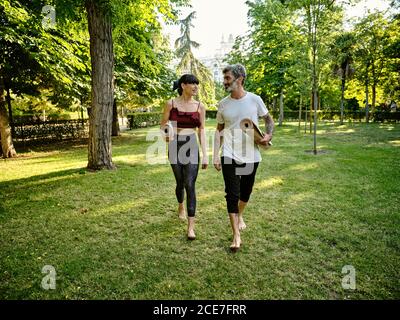 Positiver tätowierte Erwachsene Mann und fröhliche Frau in legerer Kleidung mit zusammengerollten Yogamatten, die sich unterhalten, während sie nach dem gemeinsamen Yoga-Üben im grünen Park spazieren Stockfoto