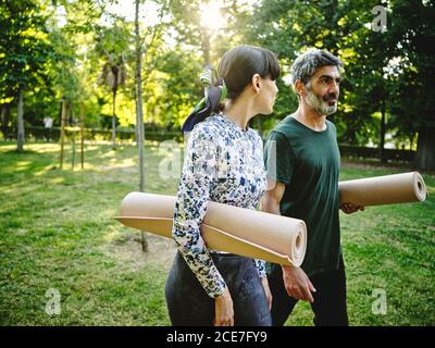 Seitenansicht eines tätowierten Mannes und einer fröhlichen Frau in legerer Kleidung mit zusammengerollten Yogamatten, die sich unterhalten, während sie nach dem gemeinsamen Yoga-Üben im grünen Park spazieren gehen Stockfoto