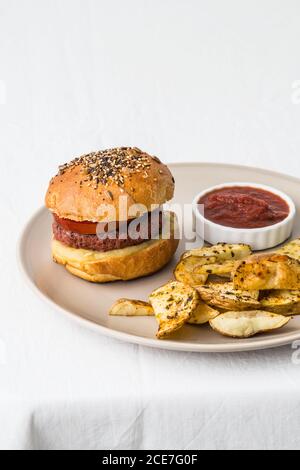 Von oben köstlicher Hamburger mit Fleischschnitzel und golden Buns mit einer Mischung aus Sesam in der Nähe knusprig geröstet dekoriert Kartoffelscheiben mit Gewürzen und Salsa Grill auf Keramikplatte Stockfoto