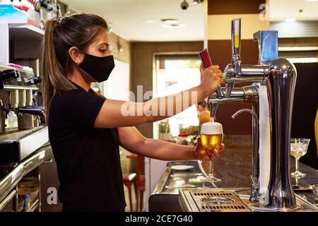 Seitenansicht des weiblichen Barkeepers in schwarzer Maske, die schaumig ausgießt Bier auf Biersäule Stockfoto