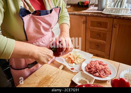 Unkenntlich beschnittene Frau auf der Schürze, die in der Küche am Tisch steht und Gemüse zum Kochen von gefüllten Paprika schneidet Stockfoto