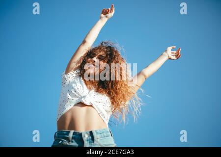 Von unten schlanke junge Frau in Casual Wear mit Lockiges Haar stehend mit erhobenen Armen und geschlossenen Augen Sommer an sonnigen Tag unter blauem Himmel Stockfoto